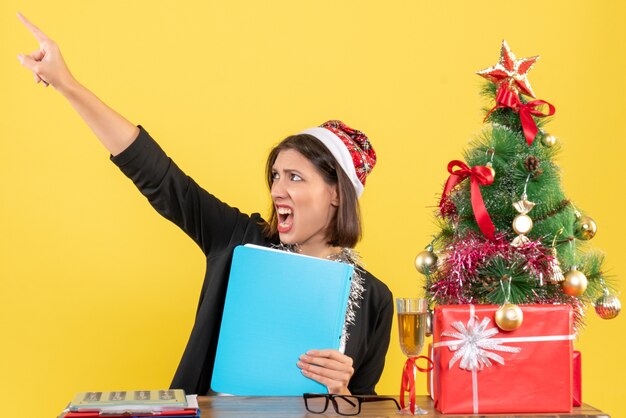 Charming lady in suit with santa claus hat and new year decorations holding document pointing up in the office on yellow isolated 