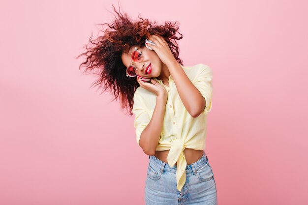 Charming lady with bronze skin and bright make-up posing with interested face expression. Good-looking mulatto woman in jeans and cotton shirt listening favorite music.