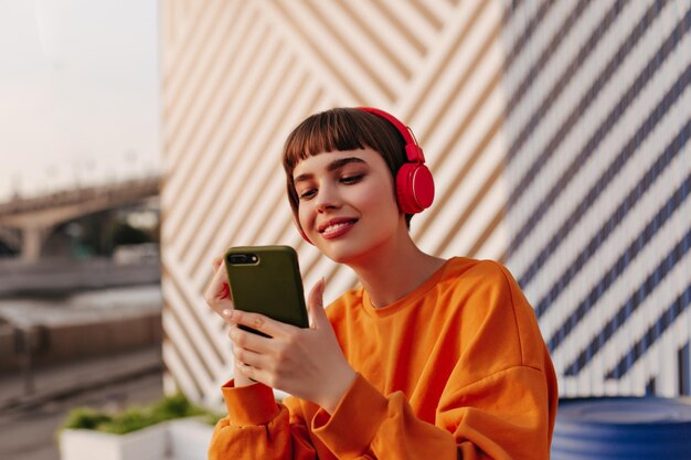 Charming lady with brunette hair in red headphones holding phone outside Girl in orange sweatshirt listening to music on striped backdrop