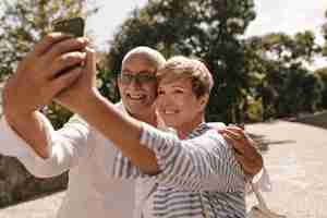 Free photo charming lady with modern blonde hairstyle in striped blouse smiling and making selfie with happy man with grey mustache in glasses and shirt outdoor