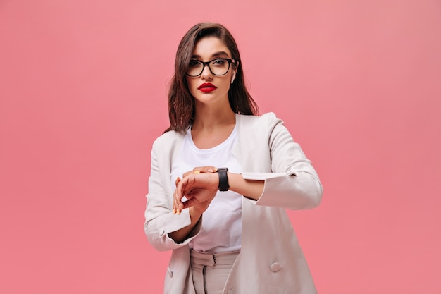 Charming lady with red lips posing on pink background.  Serious young woman in white suit and glasses looks at camera on isolated backdrop.