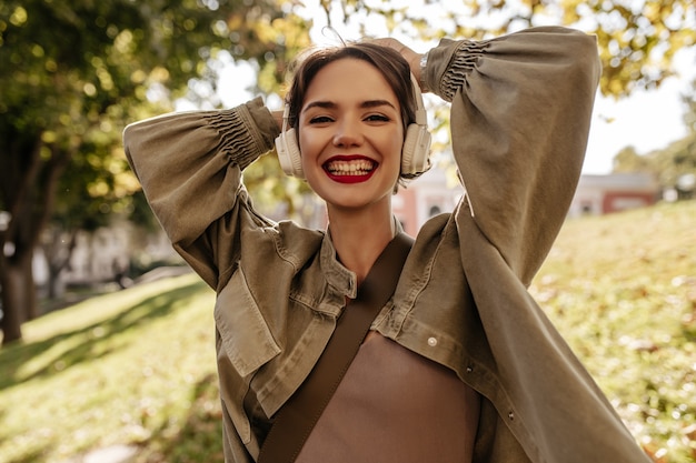 屋外で真摯に笑うデニム長袖ジャケットのショートヘアのチャーミングな女性。明るい唇のヘッドフォンで若い女性が外でポーズします。