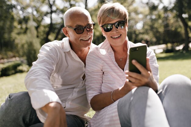 Charming lady with short hairstyle in sunglasses and pink blouse looking into smartphone and smiling with grey haired man in white shirt in park