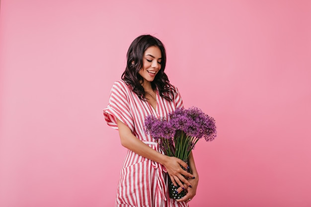 Очаровательная латинская женщина с любовью смотрит на букет фиолетовых полевых цветов. девушка в полосатом платье смущенно позирует.