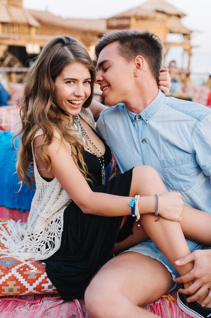 Charming laughing curly girl in elegant dress and trendy accessories sitting next to her boyfriend and posing on blur background