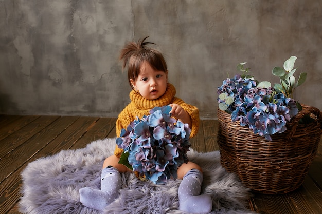 Free photo charming little baby-girl in orange sweater explores blue hydrangeas sitting on warm blanket