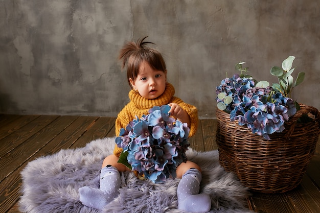 Charming little baby-girl in orange sweater explores blue hydrangeas sitting on warm blanket