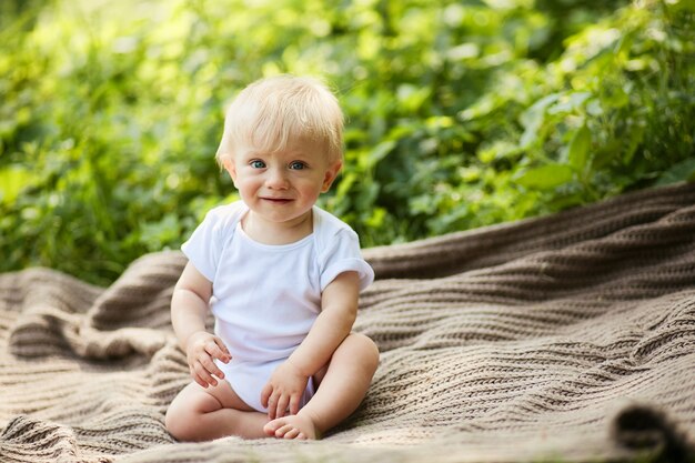 Charming little blonde boy has fun sitting on the plaid in a summer park 