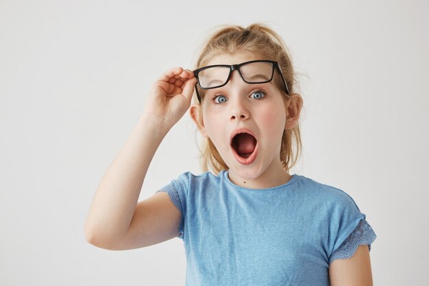 Charming little blonde lady with blue eyes reises glasses with hand in happy excitement of new big bed in her sleeping room as a present for her birthday.