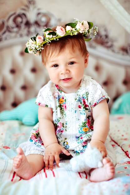 Charming little girl in wreath of roses sits on soft blankets