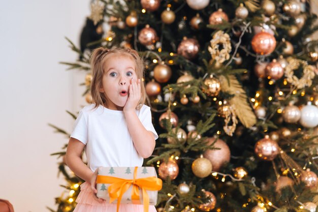 매력적인 어린 소녀가 크리스마스 나무 배경에 선물을 들고 있다