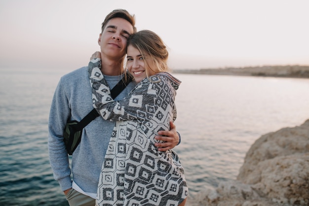 Free photo charming long-haired girl posing with her stylish boyfriend in trendy hat, gently touching his shoulder. portrait of beautiful loving couple in cute outfit sitting on the road at summer evening