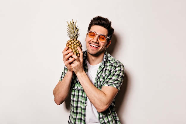 Charming man in checkered outfit and orange glasses smiles sweetly and holds pineapple.
