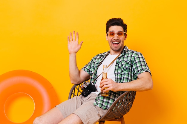 Free photo charming man in green plaid shirt waving his hand, laughing, holding bottle of beer and retro camera on orange space.