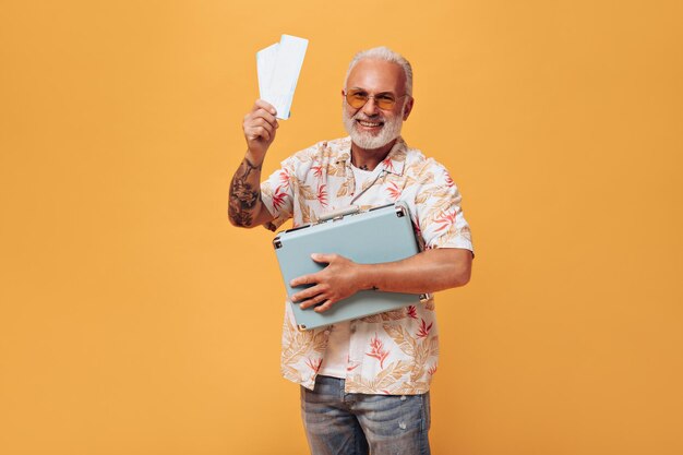 Charming man in plant print shirt poses with tickets and suitcase on orange background Grayhaired guy with beard in summer clothes is having fun