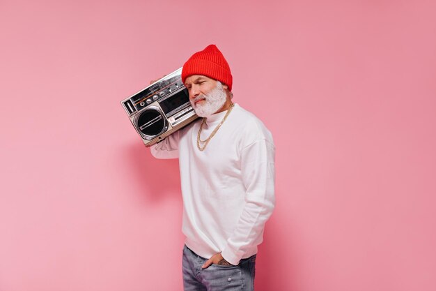 Free photo charming man in stylish hat posing with record player on pink background grey bearded adult guy in orange hat with gold chains smiling