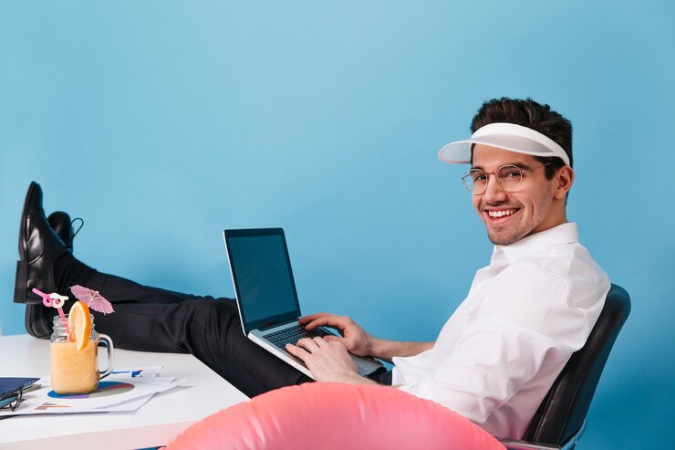 Charming man in white shirt, cap and glasses is against blue space. Guy holds laptop and works on vacation.