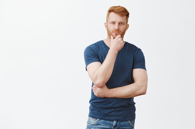 Charming masculine redhead man in blue t-shirt, touching beard and gazing with thoughtful look, thinking, making decision or choice over gray wall, having interesting idea in mind