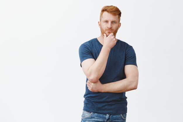 Charming masculine redhead man in blue t-shirt, touching beard and gazing with thoughtful look, thinking, making decision or choice over gray wall, having interesting idea in mind