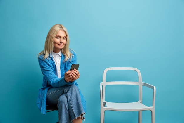 Free photo charming middle aged woman sits in queue near cabinet poses on chair against blue wall, uses smartphone