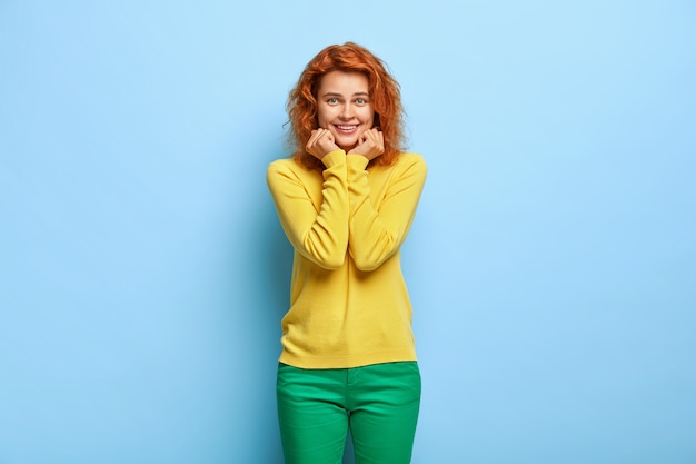 Charming millennial woman with wavy red hair posing against the blue wall