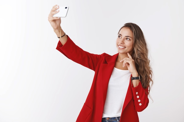 Очаровательная современная женщина-офисный служащий делает селфи, публикуя онлайн-блог, рассказывая подписчикам о новой работе, протягивая руку, держащую смартфон, фотографируя себя улыбаясь экран дисплея