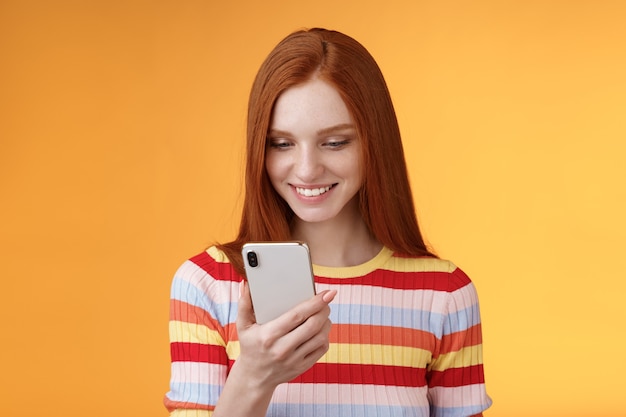 Очаровательная современная рыжая студентка колледжа, проверяющая окно сообщения со смартфоном, выглядит счастливым, улыбающимся, довольный дисплей мобильного телефона получает сотню лайков, фото пост онлайн, оранжевый фон.