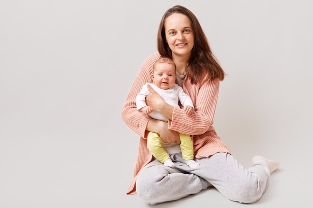 Charming mother holding newborn baby and looking directly at front with pleasant smile