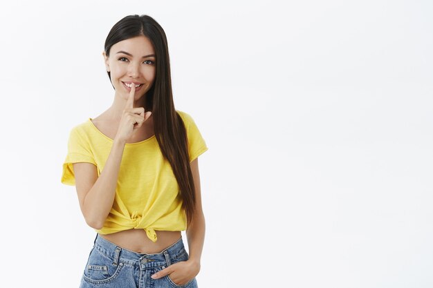 Charming mysterious and flirty young woman in trendy yellow t-shirt making shush gesture with index finger