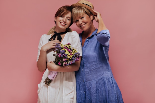 Charming old woman with short blonde hair in blue dress and straw hat smiling with girl in white clothes with beautiful flowers on pink background.