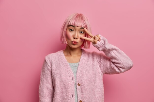 魅力的なピンクの髪の少女は勝利のジェスチャーを示し、顔に指でピースサインを作り、優しい自信を持って表現し、暖かいセーターを着ています
