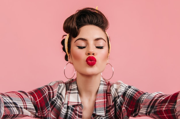 Charming pinup girl posing with kissing face expression. Studio shot of carefree young woman taking selfie on pink background.