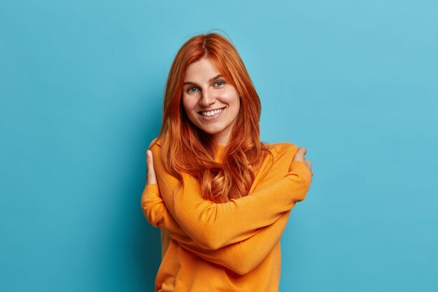 Charming pleased redhead European woman embraces own body dressed in casual orange jumper smiles pleasantly feels comfortable.