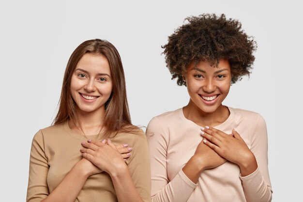 Free photo charming pretty females with pleasant smile, hold palms on heart in grateful gesture, being touched to recieve expensive gift from parents, model against white wall. gratitude concept.