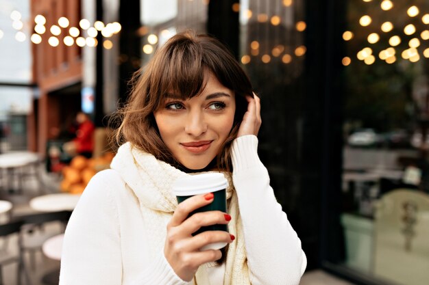 Charming pretty lady dressed white sweater and scarf drinking coffee outside on lights background High quality photo