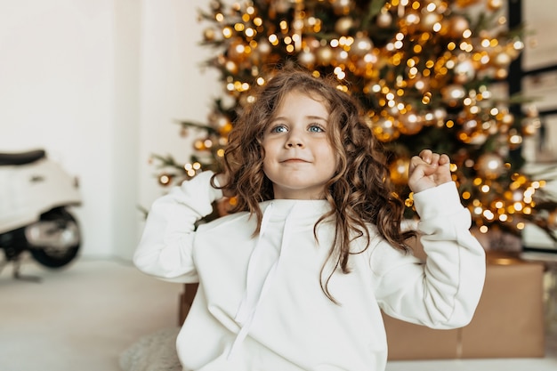 無料写真 ライトが付いているクリスマスツリーの前で笑っている白い服のカールを持つ魅力的なかわいい女の子