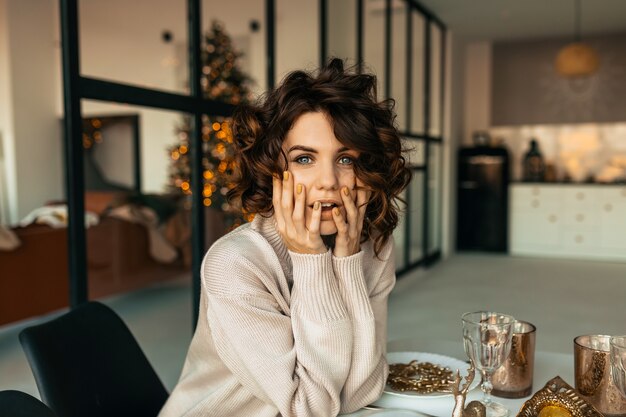 Charming pretty woman with shirt wavy hairstyle posing with surprised emotions while sitting at Christmas table over Christmas tree