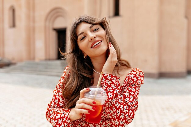 Charming pretty woman with wavy hair wearing bright summer dress walking outside with orange summer drink and smiling at camera Outdoor portrait of happy stylish girl in the city