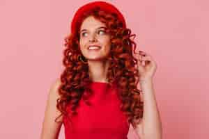 Foto gratuita affascinante ragazza dai capelli rossi con un sorriso distoglie lo sguardo. donna in cappello e top rosso tocca i capelli.