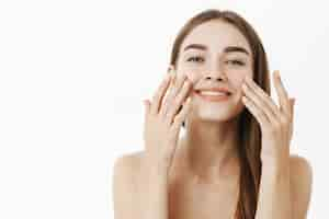 Free photo charming relaxed and gentle young woman making cosmetological procedure applying facial cream on face with fingers and smiling broadly feeling perfect, taking care of skin