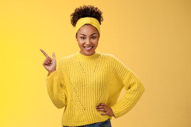 왼쪽 상단을 가리키는 스웨터를 입은 매력적이고 세련된 아프리카계 미국인 소녀는 자신감 넘치는 활기찬 미소 노란색 배경에 서서 최고의 선택 모양의 카메라를 보여주는 상품을 홍보합니다.