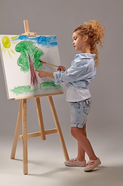 Free photo charming school girl whith a curly blond hair, wearing in a blue shirt, white t-shirt and blue jeans shorts is painting with a watercolor brush on an easel, standing on a gray background.