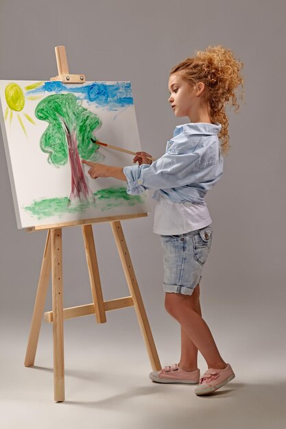 charming school girl whith a curly blond hair, wearing in a blue shirt, white t-shirt and blue jeans shorts is painting with a watercolor brush on an easel, standing on a gray background.