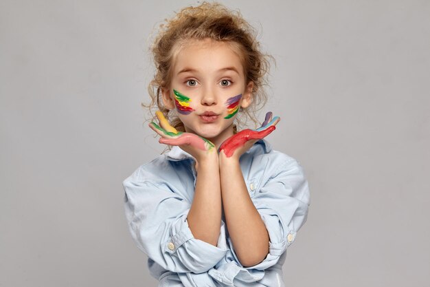 Charming schoolgirl having a brush in her chic curly blond hair, wearing in a blue shirt and white t-shirt. She has painted her cheeks and giving a kiss at the camera on a gray background.