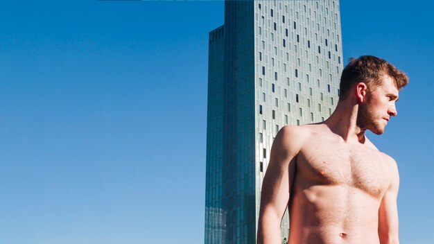 현대적인 건물 앞에 서있는 매력적인 shirtless 남자 멀리보고