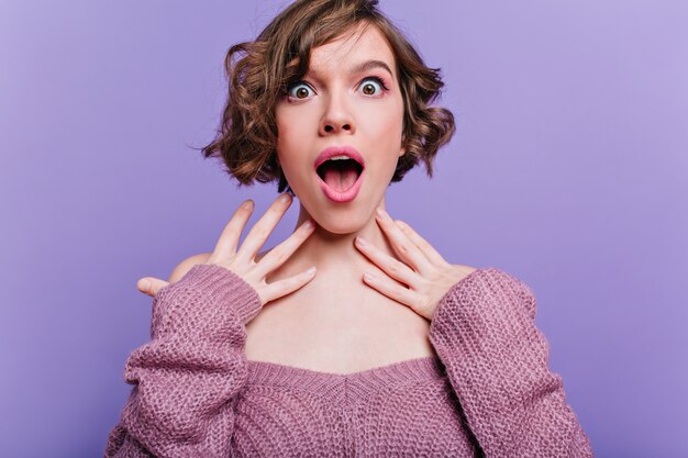 Charming shocked girl with big dark eyes posing on purple wall. Spectacular young lady with short curly hairstyle expressing amazement.