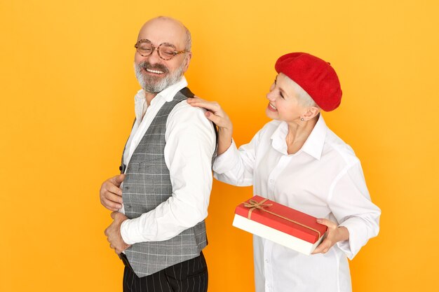 Charming short haired middle aged woman in red bonnet holding box of chocolate giving birthday present to her handsome cheerful husband