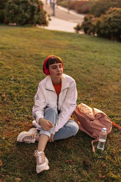 Charming shorthaired woman in jeans and white jacket sits on grass Cool girl in stylish outfit listens to music in red headphones outdoors