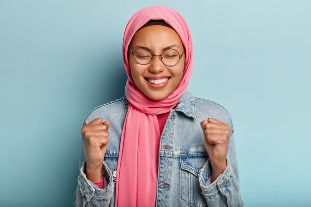 Очаровательная улыбающаяся женщина носит традиционную арабскую вуаль, сжимает кулаки, празднует достижения, приветствует победу
