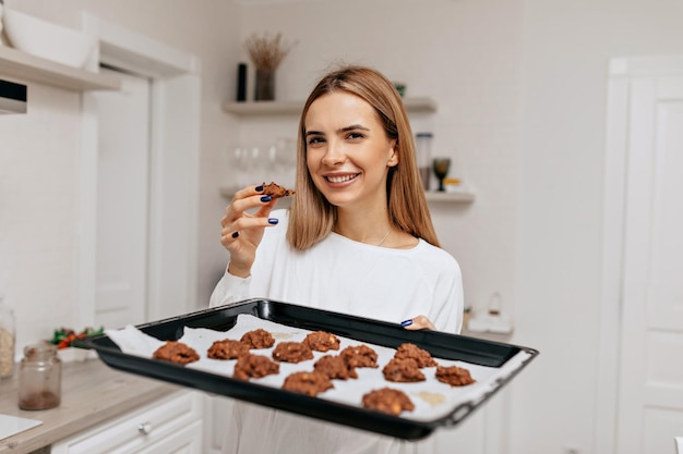 Очаровательная улыбающаяся девушка со счастливой улыбкой держит противень с печеньем и позирует перед камерой на домашней кухне